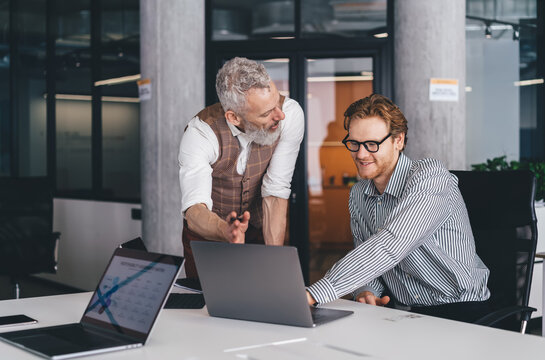 Business mentor stands beside younger colleague, smiling while discussing laptop screen. Highlights cooperative leadership, tech collaboration, and intergenerational digital teamwork.