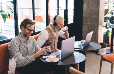 Young man smiles at smartphone while coworker sips coffee beside laptop. Friendly energy and mobile focus reflect relaxed yet productive digital culture in cafe-based freelance routine.