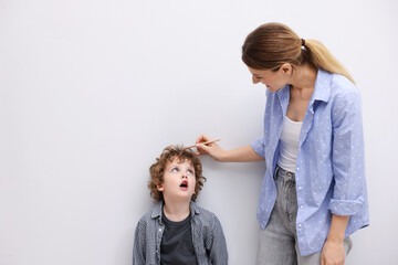 Mother measuring her little son’s height near white wall indoors. Space for text
