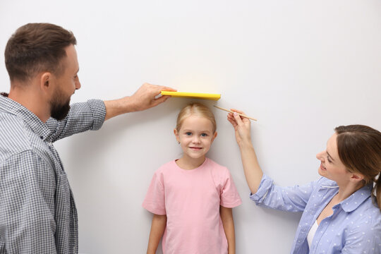 Parents measuring their little daughter’s height near white wall indoors