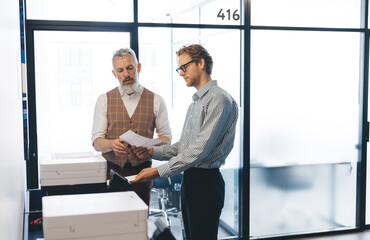 Fototapeta premium Older businessman explains printed documents to younger colleague near printers. Scene symbolizes hands-on leadership, team guidance, and productive tech-document integration in workflow.