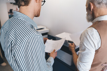 Close-up of two male colleagues handling printed reports at the printer. Represents tangible output...