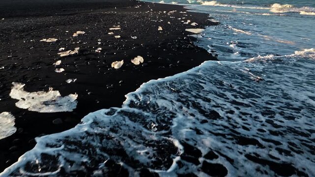 ce chunks on Diamond Beach Jokulsarlon Iceland. Drone captures waves washing over glacial ice on the black sand shore during golden light.