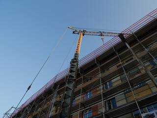 Construction crane operating beside modern building under renovation with metal scaffolding and clear blue sky, symbol of urban development, industrial growth and architectural progress in contemporar