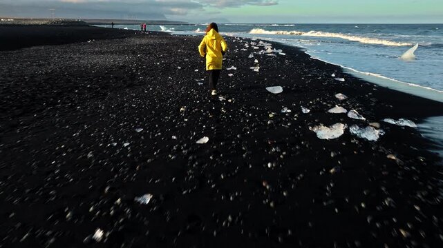 Woman in yellow jacket walking on Diamond Beach Iceland. The drone follows her walking along ice-filled shoreline under soft sunlight.