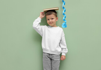 Cute little boy with book on his head near pale green background. Measuring tape handing beside him