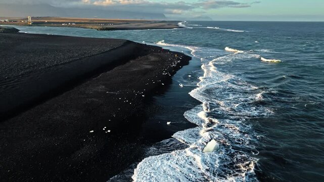 Aerial of coastline Diamond Beach Iceland. The drone moves above ocean waves meeting black volcanic sand and floating ice.