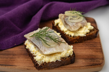 A piece of herring with dill on black bread. Two sandwiches on a wooden board with a towel.