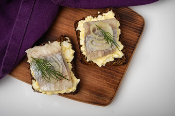 A piece of herring with dill on black bread. Two sandwiches on a wooden board with a towel.