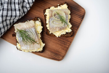 A piece of herring with dill on black bread. Two sandwiches smorebrode on a wooden board with a towel.