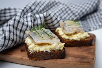 A piece of herring with dill on black bread. Two sandwiches on a wooden board with a towel.