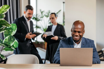 African American man in a blue suit is working on a laptop at a modern office desk, with colleagues discussing documents and greenery in the background, showcasing teamwork and productivity