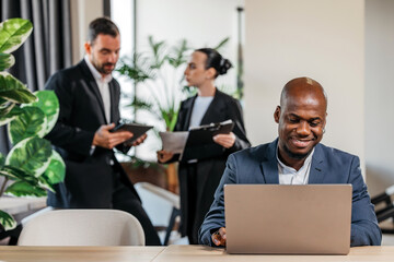 African American man working on laptop at modern office desk, while colleagues discuss project in background, showcasing teamwork and collaboration in a professional environment