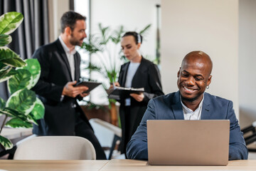 African American man working on laptop in modern office, surrounded by colleagues discussing project details, showcasing teamwork and collaboration in a professional environment