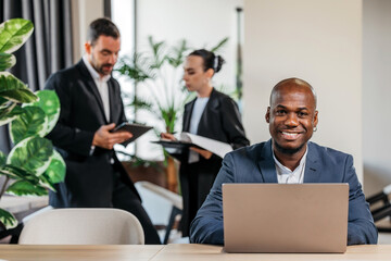 African American man in a business suit is smiling at a laptop in a modern office, with colleagues discussing documents in the background, showcasing teamwork and professionalism