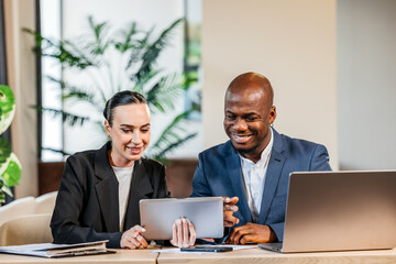 Professional african american man and woman collaborating at a modern workspace, reviewing data on a tablet, surrounded by greenery and a laptop, showcasing teamwork and innovation