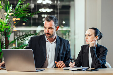 Business professionals engaged in a collaborative discussion, analyzing data on a laptop, surrounded by modern office decor and greenery, showcasing teamwork and productivity