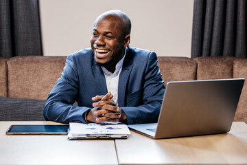 African American businessman in a blue suit, smiling confidently at a desk with a laptop and tablet, showcasing a positive atmosphere in a modern business meeting