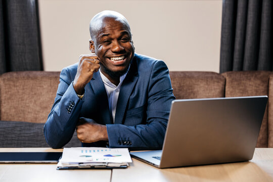 African American businessman in a blue suit celebrates success at a business meeting, seated at a desk with a laptop and documents, conveying a positive professional atmosphere - Powered by Adobe