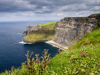 View from a cliff of Moher walking trial in rough Irish nature and blue ocean surface and cloudy sky. Famous landmark in Ireland. Travel and tourism. Beautiful Irish nature.