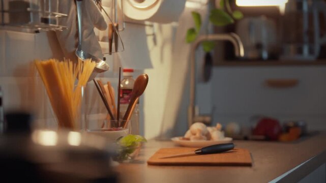 A chef prepares dinner in a warm kitchen filled with ingredients and utensils. The evening light creates a welcoming atmosphere, highlighting the organized workspace where pasta and vegetables await