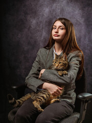 Dark and moody business portrait of a young business woman holding cute tabby cat, grey painted background. The model is in suit and sitting on a chair and has long hair and slim body type.