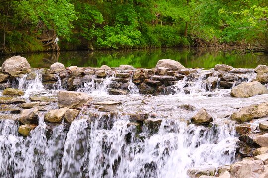 A waterfall cascading over a series of rocks.white and foamy water flows over the ledges with lush green tree background. Side view nature photo. Prairie creek park in Richardson, Texas, USA.
