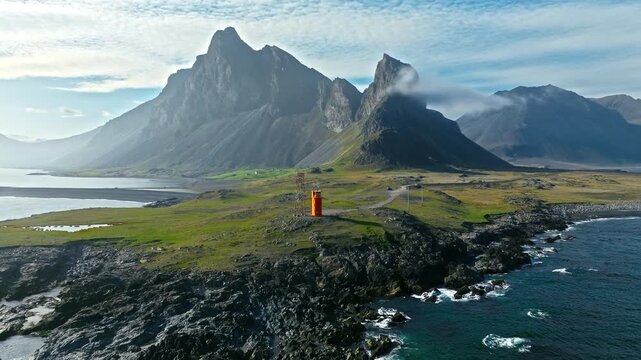 Drone flying over Hvalnes lighthouse Iceland. Smooth motion reveals bright orange tower with mountain backdrop and ocean horizon.