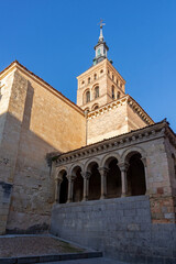The Romanesque-Mudéjar brick tower and arched portico of the Church of San Martín are bathed in warm, late afternoon sunlight, set against a bright blue sky in Segovia