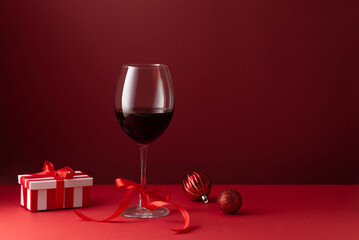 Elegant still life composition with a glass of red wine, Christmas ornaments, and a striped gift box tied with ribbon on a deep red background