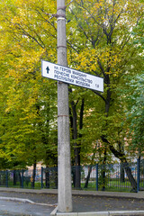 Street sign on a concrete post indicating directions to Heroes of Maidan Square and the Honorary Consulate of the Republic of Moldova, surrounded by autumn trees and urban fencing.