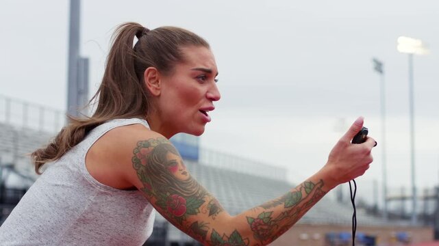 Female athletic coach watches a stop watch as her students run a cardio drill on a track outdoors.