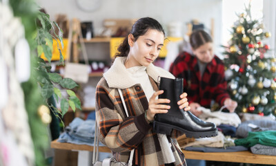 Armenian woman in a warm jacket with a fur collar chooses black platform boots against the background of a Christmas tree in a store. Shoppers on New Year sale
