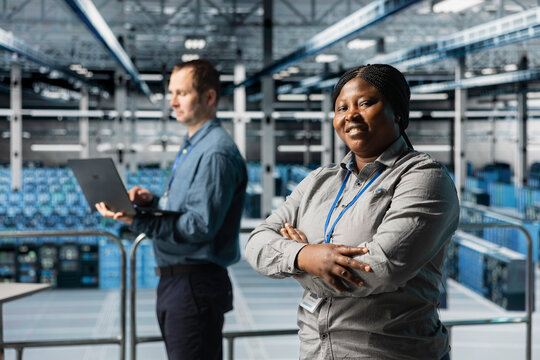 Portrait of smiling server room engineer next to coworker troubleshooting errors using software on laptop. Cheerful african american woman in data center and colleague fixing bugs using notebook - Powered by Adobe