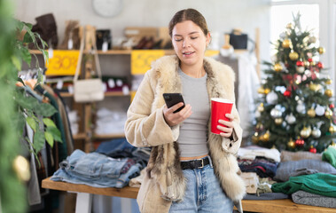 Glad young girl with paper cup in hand scrolling mobile phone during Christmas shopping