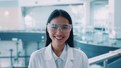 A smiling Asian woman in a lab coat and safety goggles stands confidently in a contemporary research facility. She represents the spirit of scientific exploration and innovation during a pandemic.
