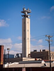 Vertical communication tower rises prominently above industrial rooftops and city utility lines against a bright blue sky.