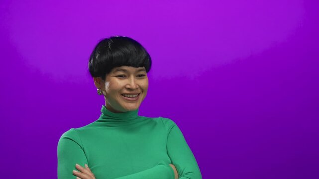 Smiling woman in green sweater against vibrant purple background exuding happiness and youthful charm with crossed arms in an isolated studio setting showcasing modern style