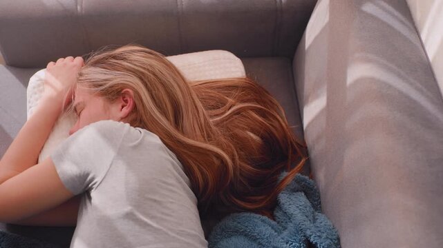 Independent woman lying sideways on couch pressing face into soft pillow resisting morning light while wrapped in blue blanket, refusing to wake up, emotionally drained