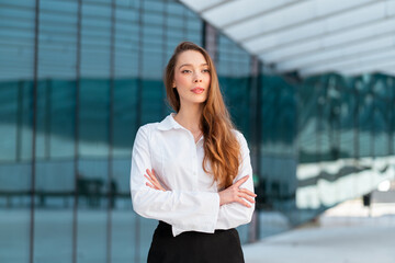 Confident businesswoman with long brown hair standing arms crossed outside modern office. Professional lady in white shirt looking away with determination
