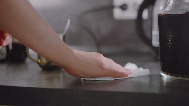Close up of human hand wiping wet kitchen countertop using paper towel beside electric kettle and coffee jar, showing household cleanliness, morning routine, personal hygiene