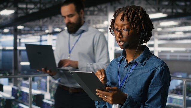 Server room admin using tablet and headset microphone to provide infrastructure tech support. Data center IT specialist resolves backend issues while coordinating with remote teams