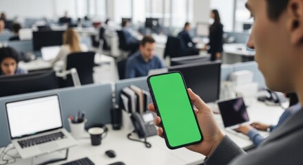 Man holding a phone with green screen in a busy office with coworkers at their desks in the background