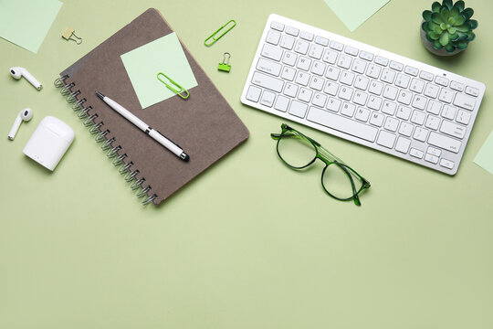 Modern office workspace with keyboard, notebook, pen, eyeglasses, and wireless earbuds on green background