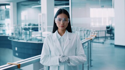 A young Asian woman in a lab coat stands confidently in a modern laboratory. She wears safety goggles and gloves, preparing for research activities related to pandemic solutions. - Powered by Adobe