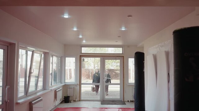 Two guys in jackets approach glass entrance of training hall, one carrying leather bag, viewed from inside room with punching bags, mats, radiators and windows, urban lifestyle moment in daylight