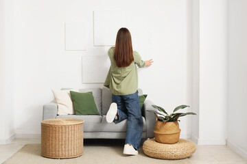 Young woman hanging picture on white wall at home, back view