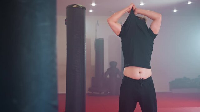 chap takes off shirt in gym environment, standing near punching bag under strong lights, showing chest and torso while preparing for workout, representing strength and determination for training