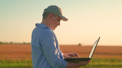 Businessman with computer in hands works in wheat field, checking harvest. Farmer in field works...