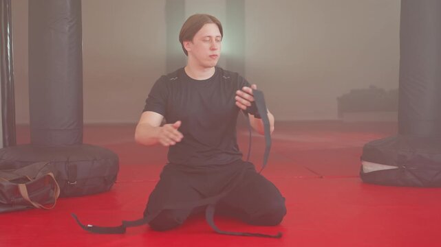 Boxer in black sportswear sits on red mat gym floor wrapping long hand bandages with focus and precision, preparing for training session with punching bags around, showing strength and readiness for